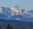 Panoramic view of snow-capped mountains and a valley with scattered trees and a body of water in the foreground.