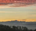 A serene view from a rooftop terrace showcasing a picturesque mountain range at sunset, with a clear sky transitioning from warm orange to deep blue hues, surrounded by silhouettes of trees and a distant forest.