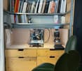 A modern, minimalist kitchen area with a coffee machine, a bookshelf filled with books, and wooden drawers.