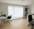 Bright, modern office with light wooden flooring, large window with white curtains, white desk with laptop and chair, bookshelf with books and decorative items, and a hanging light fixture.