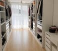 A well-organized walk-in closet with built-in white wardrobes, shelves, and drawers. The space features light wooden flooring, large windows with white curtains, and various clothing and shoes neatly arranged.