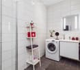 Modern bathroom with white tiles, a sink with a mirror, a washing machine, a shelf with towels and items, and a small plant on the counter.