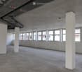 Empty room with white walls, concrete floor, large windows, and visible structural beams and pipes.