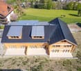 A modern two-story house with a dark grey metal roof, solar panels, and large wooden-framed windows. The house features a spacious patio area with a tiled floor and is surrounded by a green field and trees.