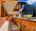 Wooden balcony with a wooden railing, overlooking a scenic mountain landscape with trees and a clear blue sky.