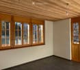 Empty room with wooden ceiling, large windows, and wooden doors, featuring a dark tiled floor and light-colored walls.