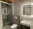 Modern bathroom with grey tiles, a glass-enclosed shower, a toilet, a sink with a mirror, and built-in lighting around the mirror.