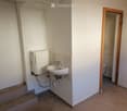 Small bathroom with a white sink, chrome faucet, and a toilet visible through an open wooden door. A water heater is mounted on the wall next to the sink.