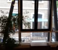 A room with large windows overlooking a brick building and a staircase. Inside, there is a wooden table, a chair, and a floor lamp near the window.