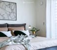 A modern bedroom with a large bed, multiple pillows, a blanket, a nightstand with a lamp, a small plant, and a large picture frame on the wall.