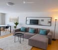 Modern living room with a grey sectional sofa, teal throw pillows, a black coffee table, a dining table with chairs, a large mirror with a TV, and potted plants.