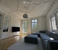 Elegant living room with ornate ceiling, large windows, grey sectional sofa, ottomans, flat-screen TV, and a modern floor lamp.