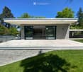 Modern two-story house with large glass windows, stone facade, and a spacious patio area surrounded by greenery.