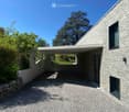 Modern stone-clad building with a flat roof and large windows, surrounded by greenery and a gravel driveway.
