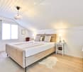 Modern bedroom with a large bed, nightstands, a chair, and a hanging pendant light. The room features light wooden flooring and white walls with a slanted ceiling.