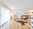 Bright living room with wooden flooring, large windows with white curtains, a grey sectional sofa, armchair, coffee table, side tables, floor lamp, decorative rug, and artwork on the walls.