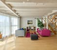 Bright living room with wooden floors, high ceilings, large windows with white curtains, two sofas, coffee table, ottoman, bookshelf, and decorative plants.