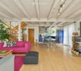 Bright living room with wooden floors, exposed wooden beams, a pink sofa, coffee table, ottoman, fireplace, dining area with a glass table and chairs, and a staircase leading to another level.
