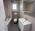 Modern bathroom with a bathtub, toilet, sink, and mirror. Features grey tiles and a window for natural light.