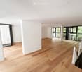 Empty room with light wooden flooring, white walls, large windows, and a staircase leading upwards.