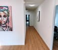Modern hallway with light wooden flooring, white walls, and recessed ceiling lighting. Features a large abstract painting on one wall and a glimpse of an office area with a desk and chair at the end.