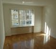 Empty room with wooden flooring, white walls, and large windows showing a view of neighboring buildings.