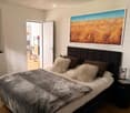 Modern bedroom with a large bed, fur throw, decorative pillows, nightstand, and a large abstract painting on the wall. An open doorway leads to a kitchen area with a table and chairs.