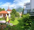 Garden with a table, chairs, umbrella, and potted plants, surrounded by greenery and a view of mountains and buildings.
