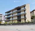 Modern multi-story building with beige facade, large windows, and balconies. Features a sleek design with glass railings and a small garden area in front.