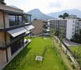 Well-maintained garden area with green grass, small trees, and a few outdoor chairs and tables near a modern building with large windows and a covered balcony.