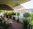 A spacious balcony with wooden decking, a large umbrella, several potted plants, a sofa, a small table, and chairs.