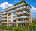 Multi-story residential building with balconies, surrounded by greenery and a garden area with outdoor furniture.