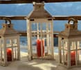 Three wooden lanterns with red candles placed on a snowy surface, likely a balcony or terrace, with a wooden railing and a blue sky in the background.