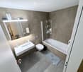 Modern bathroom with a white sink, toilet, bathtub, and large mirror. Grey tiled walls and floor, with a small shelf holding toiletries.