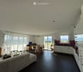 Open-plan living and dining area with a sofa, coffee table, dining table, chairs, and a modern kitchen with cabinets, refrigerator, and oven. Large windows offer a scenic view.