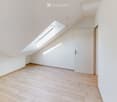 Empty attic room with wooden flooring, white walls, and a skylight window allowing natural light to enter. The room features a single white door and a textured wall on one side.