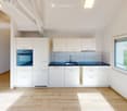 Modern kitchen with white cabinets, a built-in oven, and a sink, featuring light wooden flooring and large windows.
