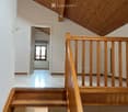 Wooden staircase with a wooden railing and a skylight above, leading to a bright, open space with white walls and a large window at the end.
