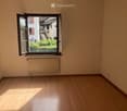Empty room with wooden flooring, white walls, a radiator, and a large window with a view of a garden and neighboring building.