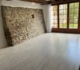Empty room with exposed wooden beams, stone wall, large windows with white curtains, and polished wooden floor.