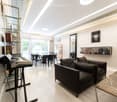 Modern, open-concept living and dining area with a sleek black sofa, dining table with chairs, a piano, and decorative mirrors. The space features a high ceiling with recessed lighting and a decorative border.