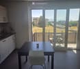 Modern kitchen-dining area with a dark wooden dining table, white chair, and a cup on the table. Large windows and sliding doors open to a balcony with outdoor furniture and a scenic view of the city and mountains.
