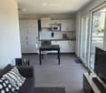Open-plan living and dining area with a grey sofa, coffee table, dining table with chairs, kitchen area with cabinets, microwave, oven, sink, and refrigerator. Large windows provide natural light.