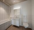 Modern bathroom with white tiles, a bathtub, a sink with a modern faucet, a toilet, and a large mirror with built-in lighting. The floor is tiled in a warm brown color.
