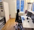 Modern office space with a large desk, ergonomic office chair, dual monitors, keyboard, mouse, and a cabinet with shelves. The room has wooden flooring and large windows offering a city view.