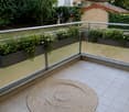 Balcony with tiled floor, glass railing, and potted plants, featuring a round woven rug in the center.