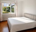 Bright bedroom with wooden flooring, large window with curtains, and a neatly made bed with a light-colored duvet and headboard.