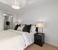Modern bedroom with a large bed, white bedding, black pillows, a nightstand with a lamp, and a decorative pendant light hanging from the ceiling.