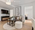 Modern living room with light wooden flooring, a grey sofa with pillows, a coffee table, a round white rug, a grey chair, a small table, a TV mounted on the wall, a cabinet, and decorative vases.