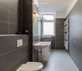 Modern bathroom with dark grey tiles, a white sink, toilet, and a window above the sink. A radiator is mounted on the wall, and a mirror is positioned above the sink.